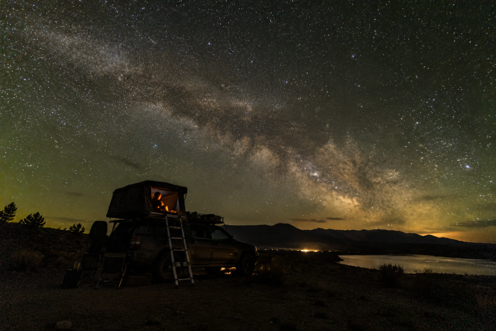 A Via Láctea sobre um acampamento à noite: um carro com uma barraca no teto iluminada por dentro, montanhas e um lago ao fundo.